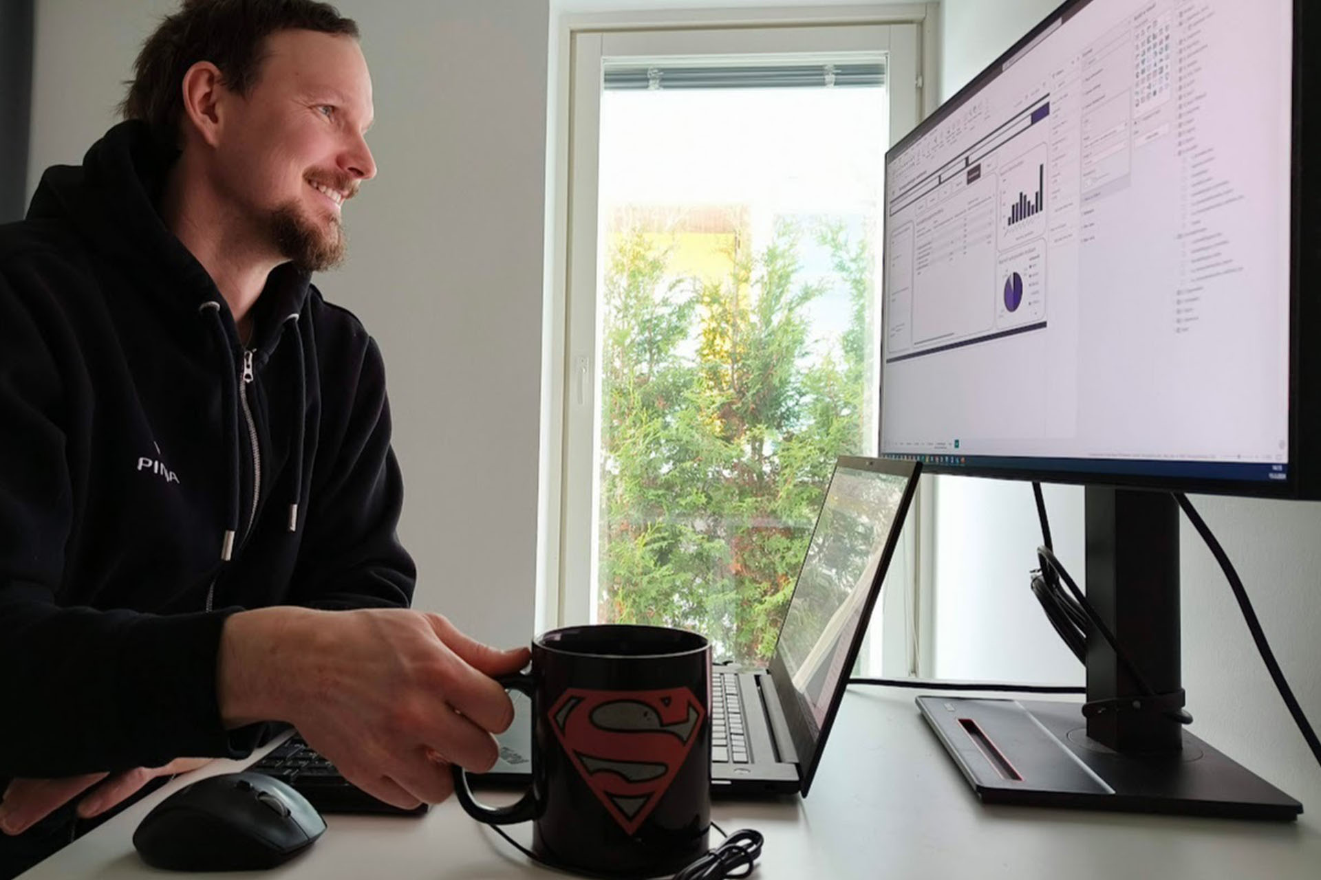 Smiling man looking at graphs on computer screen holding superman coffee cup