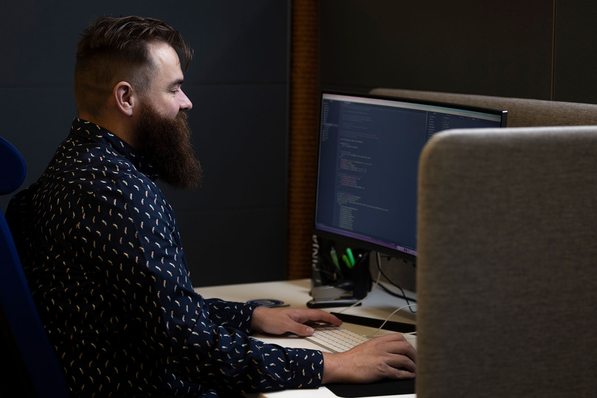 Siniseen kauluspaitaan pukeutunut parrakas mies istuu työpöydän ääressä edessään koodia ruudulla