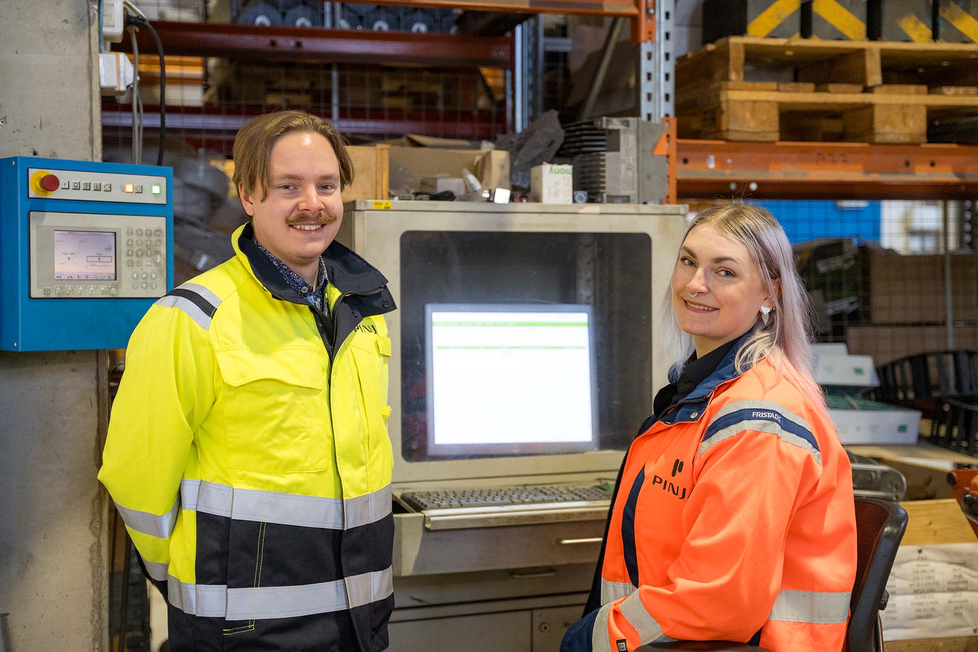 A smiling man and woman in safety equipment in the production facilities in front of the screen