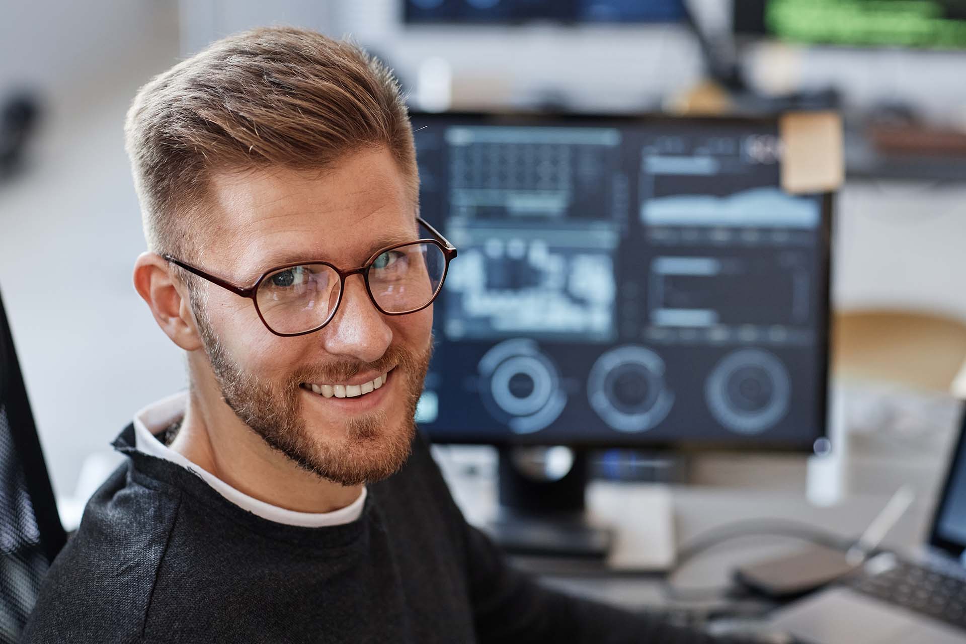 A bespectacled man smiles at the camera with a screen showing analytics in the background
