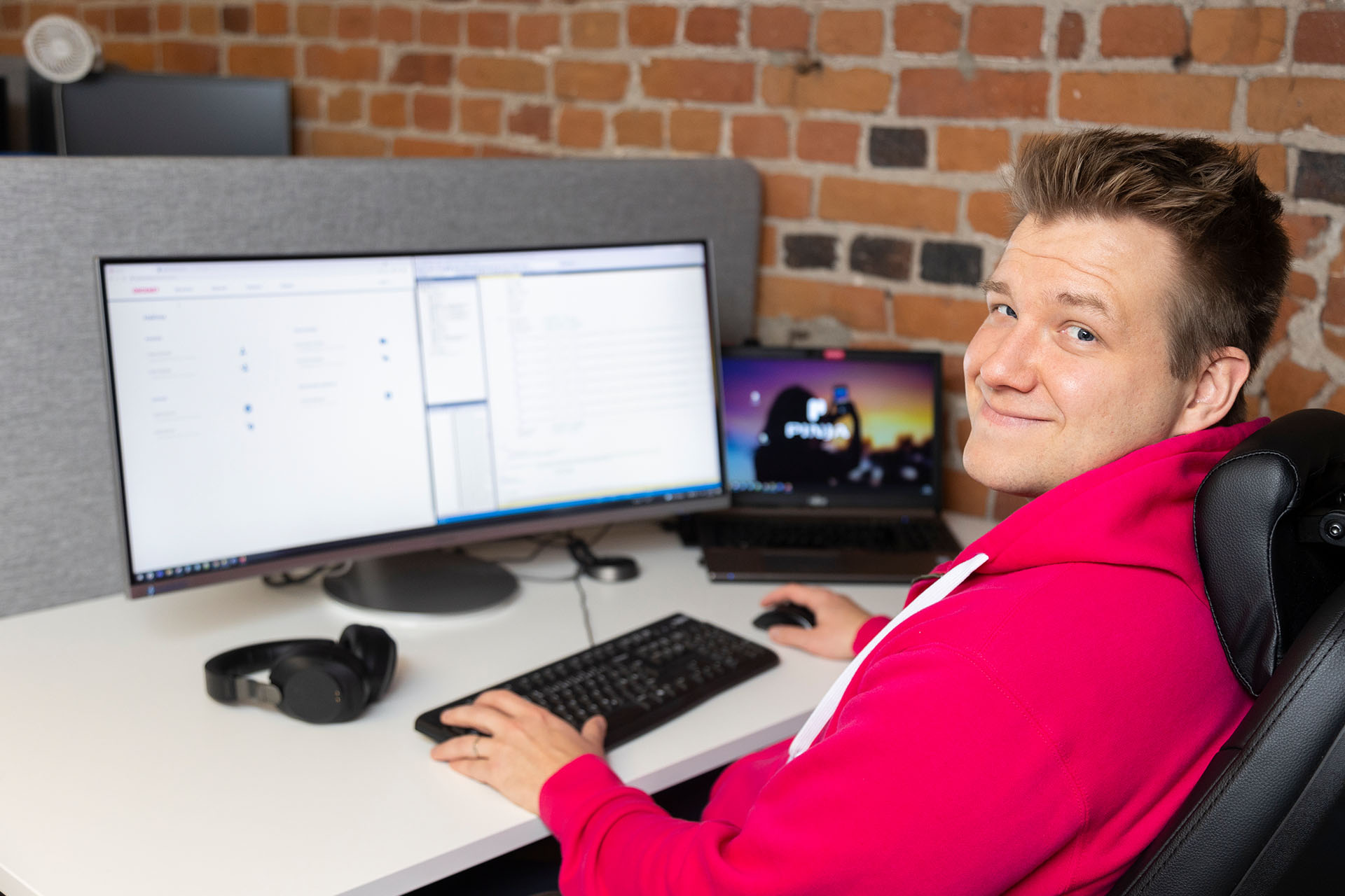 A man in a pink hoodie sitting at a computer looks over his shoulder at the camera, smiling