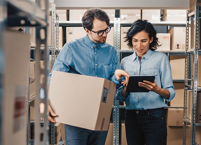 A woman showing a man holding a cardboard box something on a tablet in a warehouse