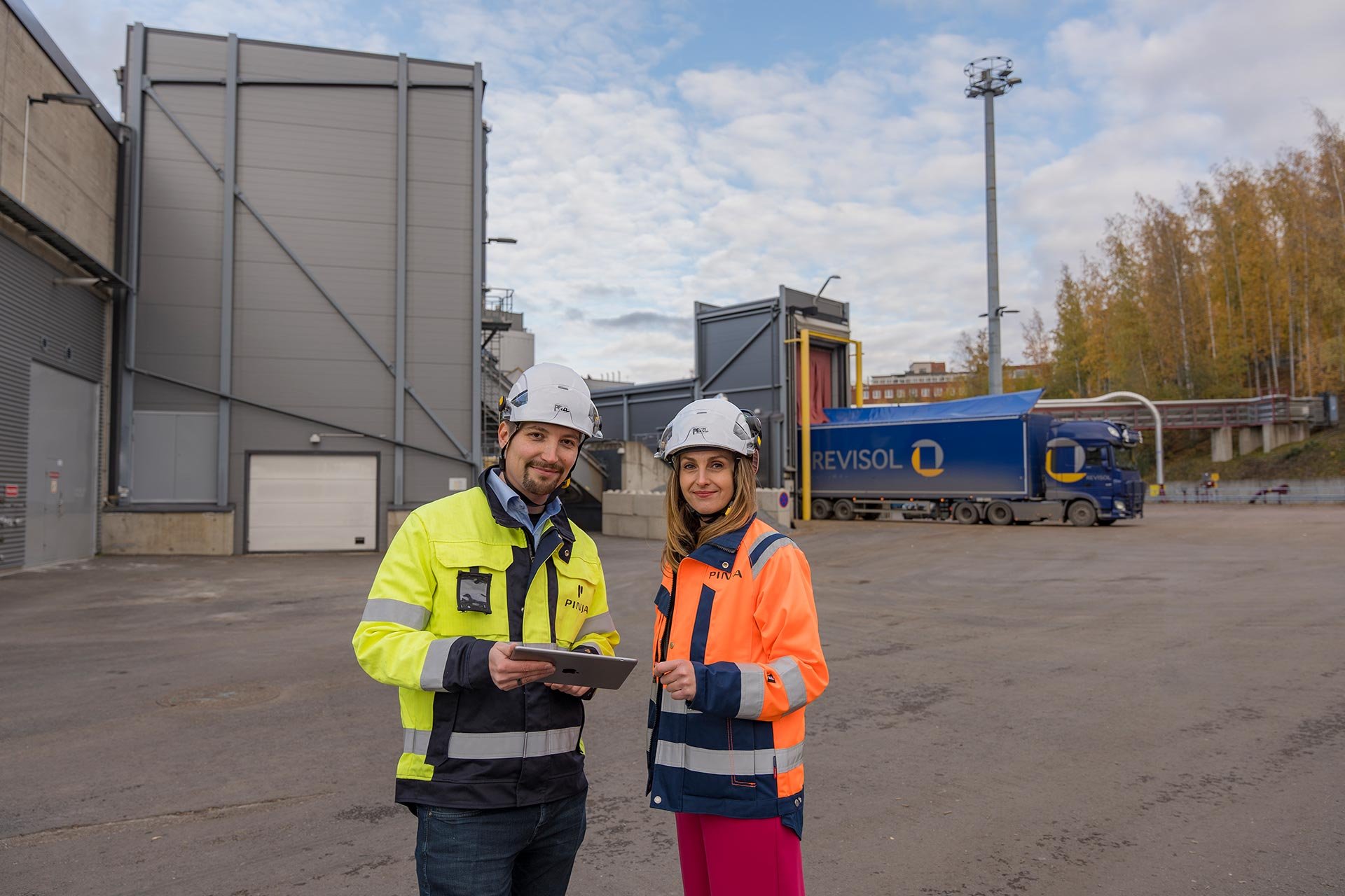 Two people wearing helmets and Pinja branded work coats outside an energy plant