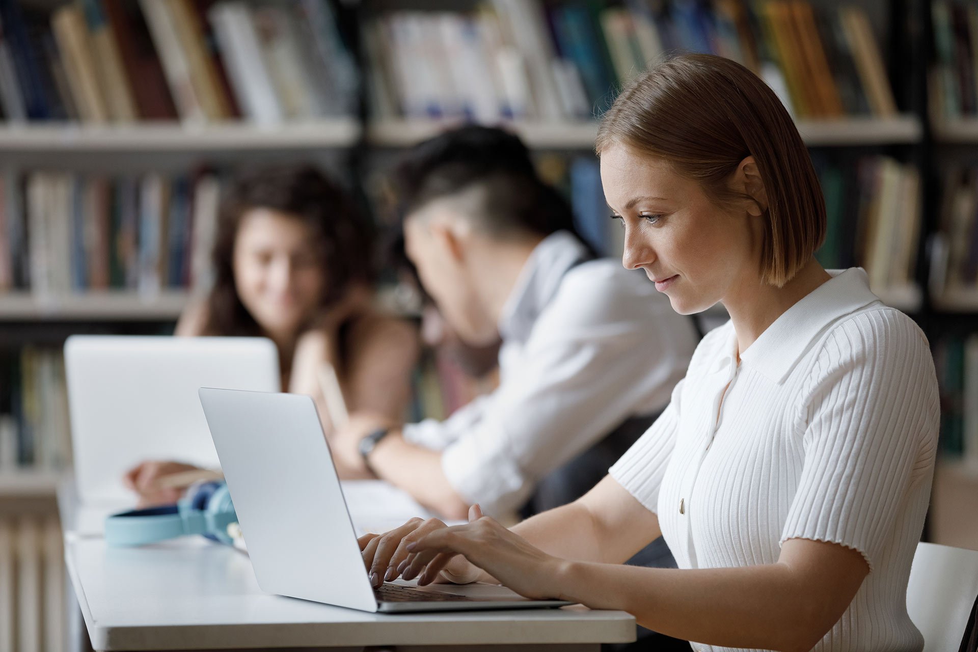 Nuori nainen työskentelee kannettavalla tietokoneella, taustalla näkyy kaksi henkilöä ja kirjahyllyjä