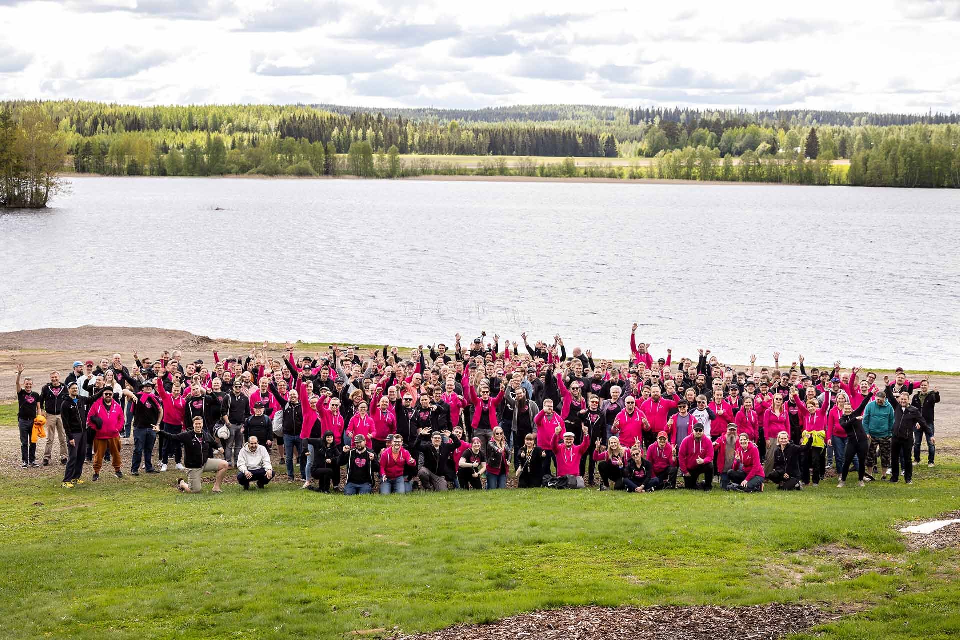 Joukko pinkkeihin ja mustiin huppareihin pukeutuneita pinjalaisia järven rannalla heiluttamassa kameraan