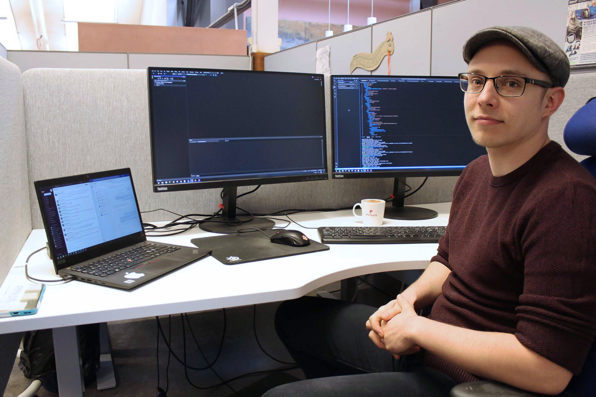 Ohjelmistokehittäjä Roni istumassa työpisteellään takanaan kaksi näytöllistä koodia